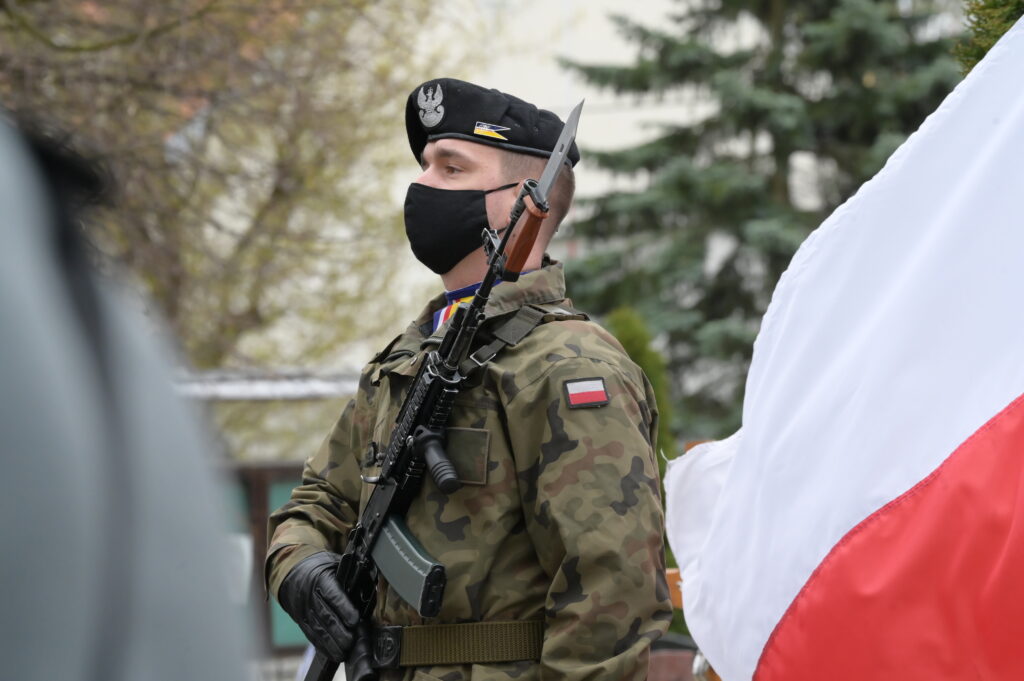 Na zdjęciu żołnierz z bronią, a przy nim powiewająca biało-czerwona flaga.