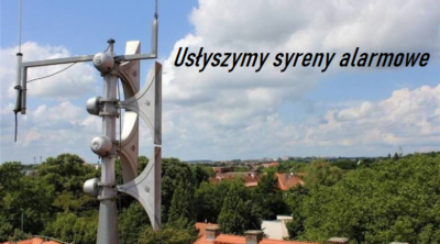 Syrena alarmowa ponad dachami domów na tle lekko zachmurzonego nieba.